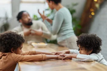 Sad African American siblings holding hands and talking at the table while their parents are arguing in the background. Copy space.